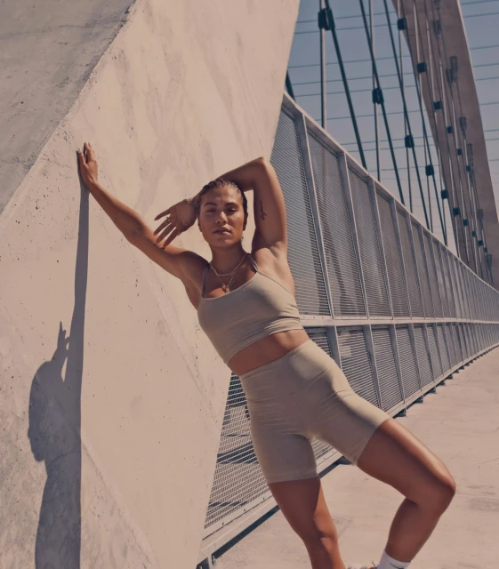 Athletic woman in tan workout gear leans against a concrete wall outside with a metal bridge in the background, capturing fitness and lifestyle.