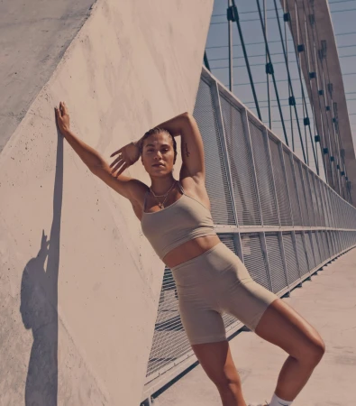 Athletic woman in tan workout gear leans against a concrete wall outside with a metal bridge in the background, capturing fitness and lifestyle.