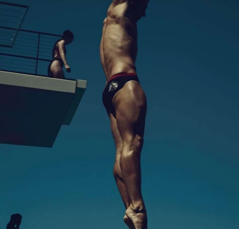 Male diver poised to jump from platform with another athlete visible on the platform against a clear blue sky, outdoor setting.