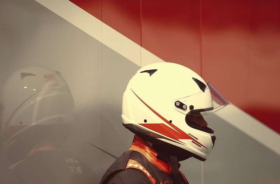 Close-up of a helmet-clad racecar driver, with a partial side view of a helmet and a red and white wall in the background.
