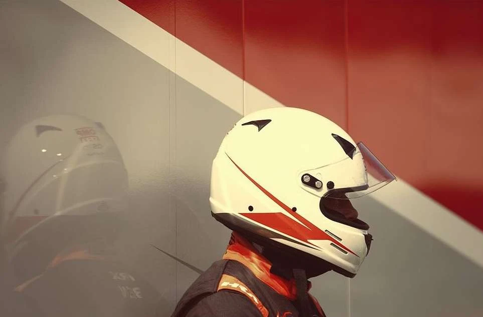 Close-up of a helmet-clad racecar driver, with a partial side view of a helmet and a red and white wall in the background.