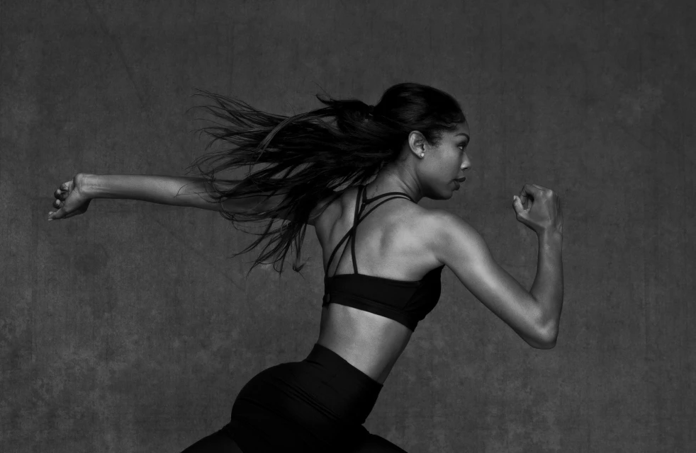 An athletic woman in black sportswear is captured mid-motion against a textured grey backdrop, her hair flowing, in a dynamic black and white studio shot.