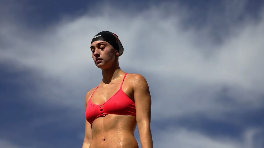 Close-up shot of a young woman wearing a swimsuit and swim cap, glistening with sweat, against a cloudy blue sky backdrop.