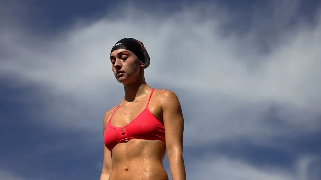 Close-up shot of a young woman wearing a swimsuit and swim cap, glistening with sweat, against a cloudy blue sky backdrop.