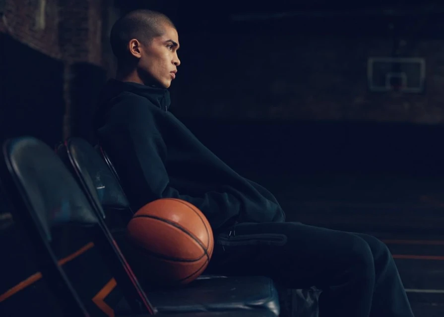 Side profile of a young adult male in dark athletic wear sitting on a folding chair with a basketball in a dimly lit indoor basketball court.