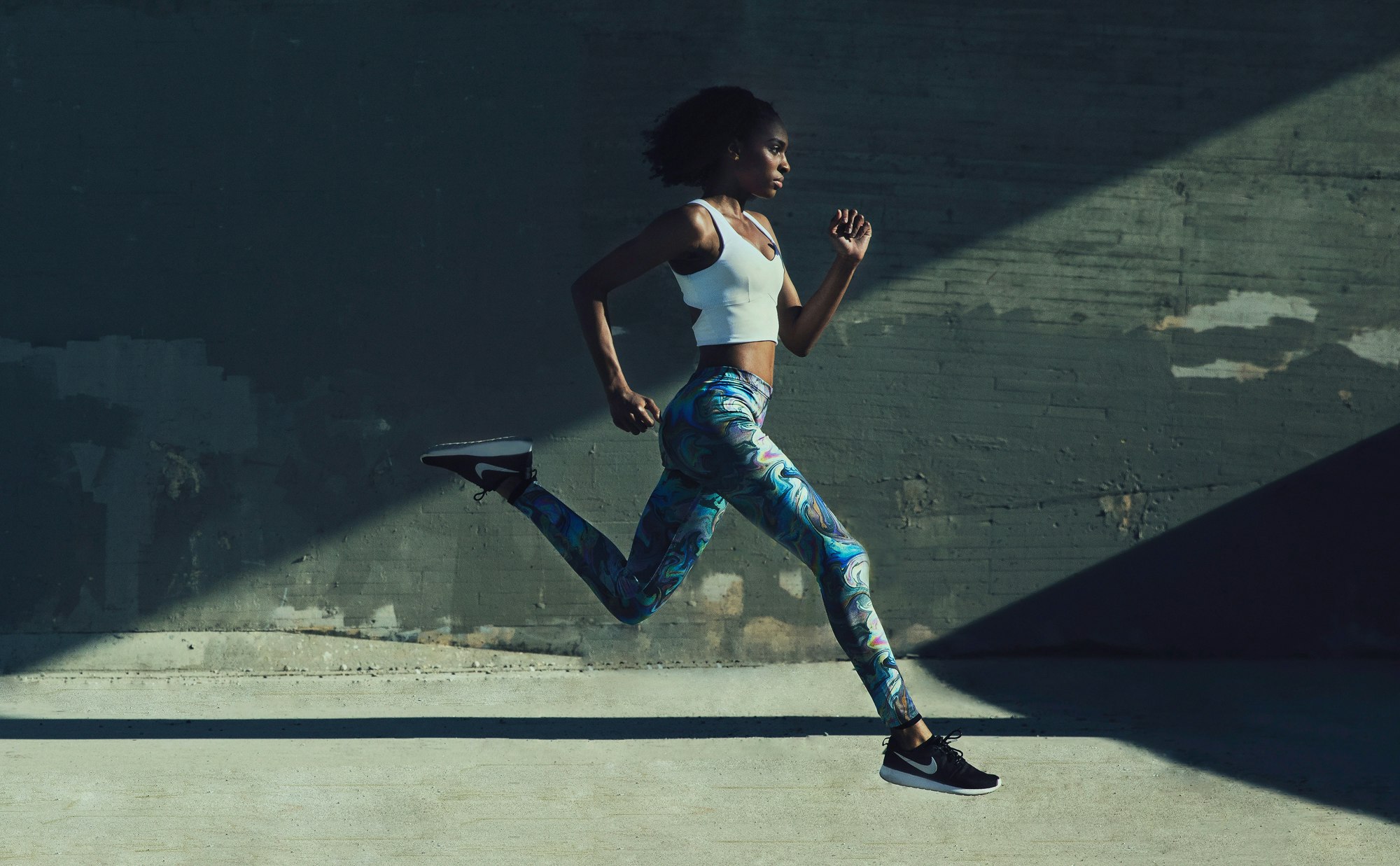 Athletic woman in activewear running outdoors, side view. Dynamic action shot with concrete wall background and directional sunlight.