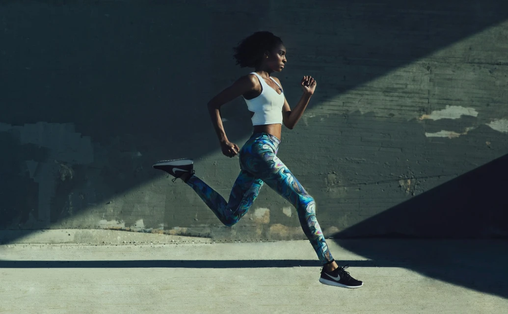 Athletic woman in activewear running outdoors, side view. Dynamic action shot with concrete wall background and directional sunlight.