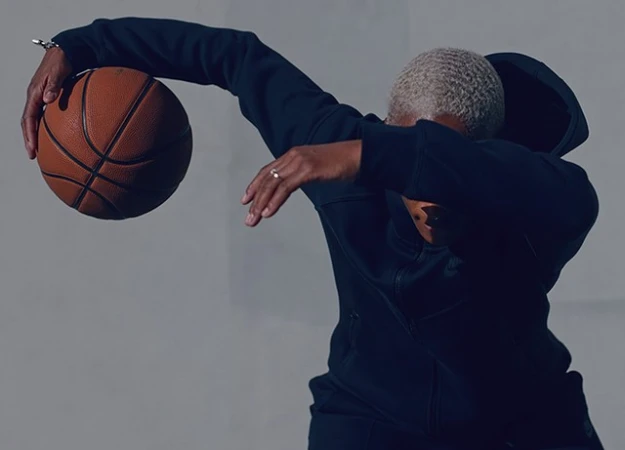 Person with short blonde hair wearing a dark zip hoodie holding a basketball against a plain grey background