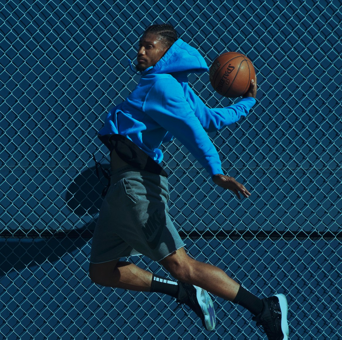 Athletic Black man in mid-air holding a basketball, wearing a blue hoodie and grey shorts, with a blue chain-link fence background.