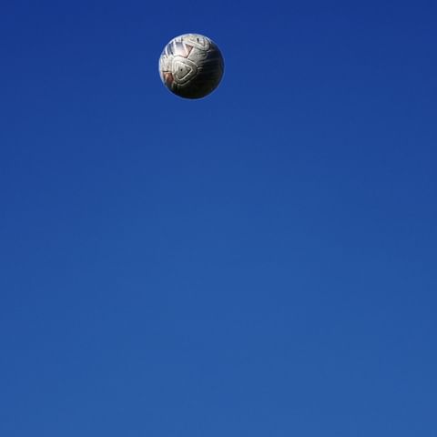 A white soccer ball is suspended in a clear blue sky with no clouds, creating a minimalist and sporty composition.