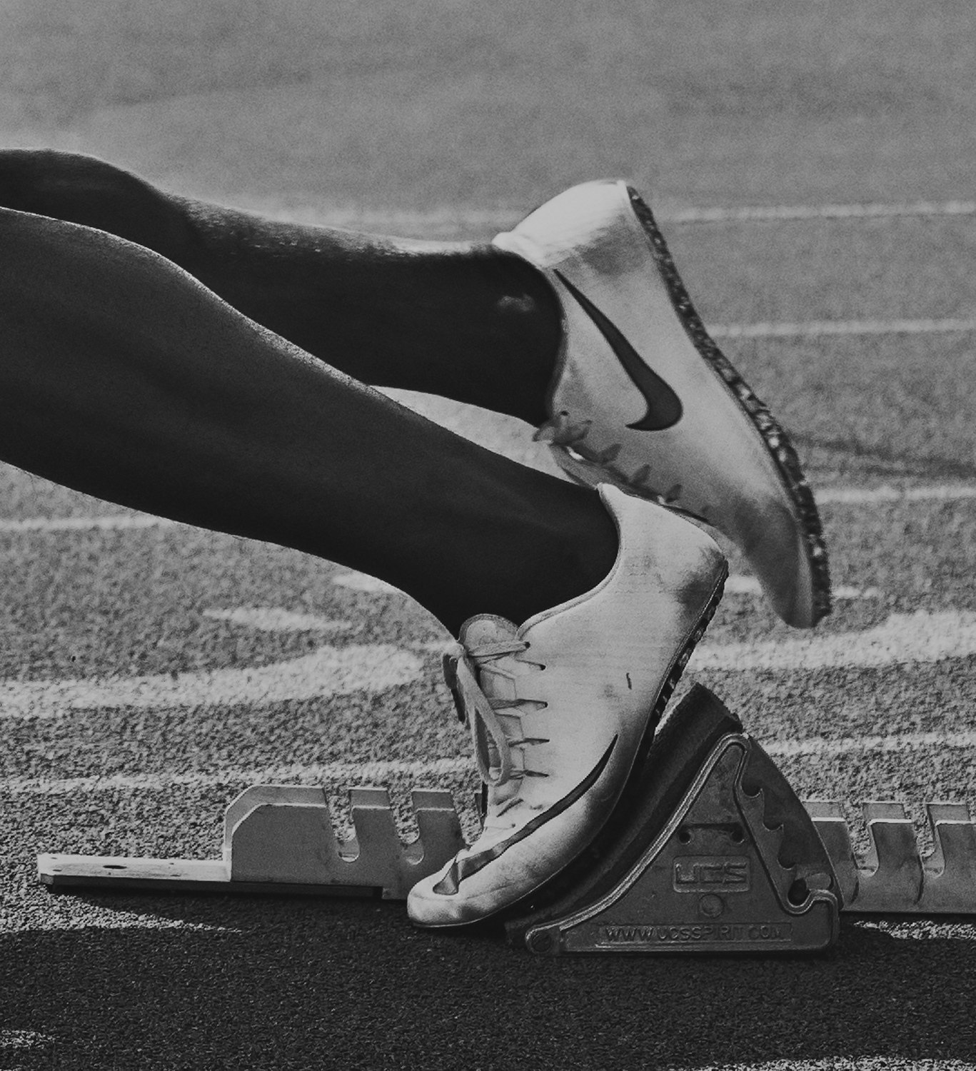 Monochrome shot of an athlete's legs and feet in Nike track shoes on a starting block, poised for a race on a textured track surface.