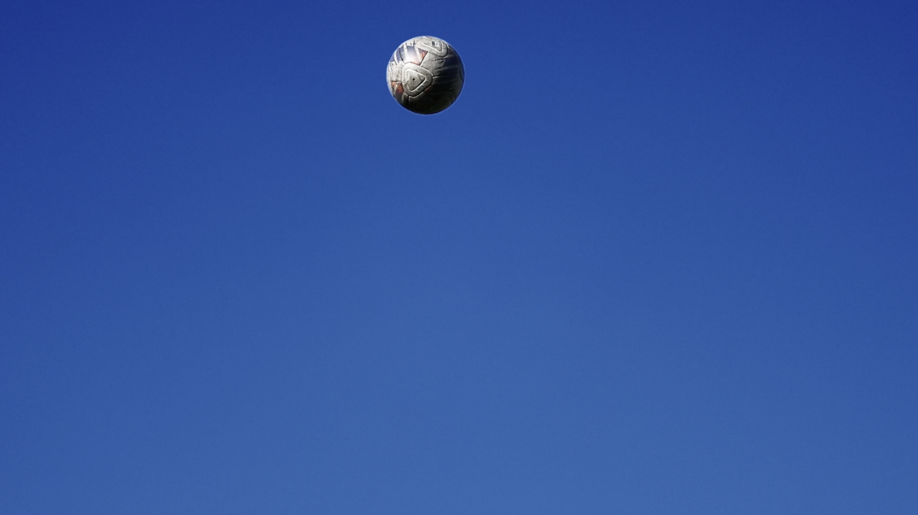 Close-up shot of a soccer ball suspended in a clear blue sky, suggesting a dynamic sports moment with a bright, sunny atmosphere.