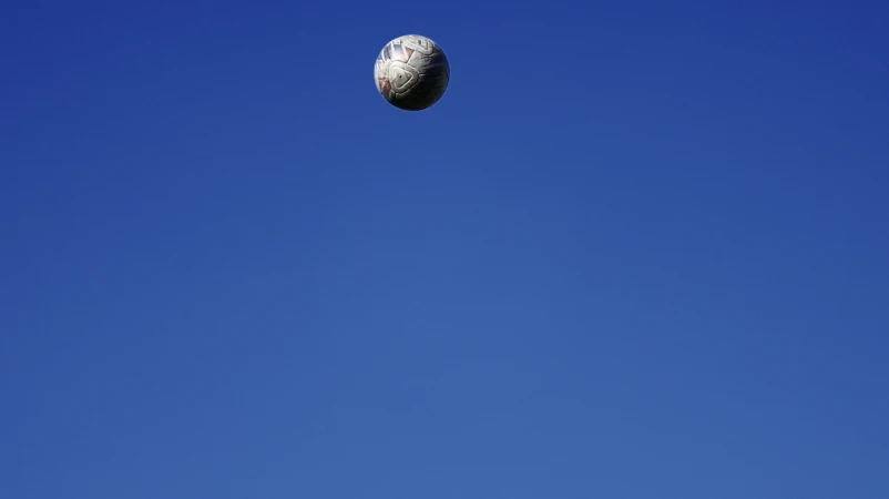 Close-up shot of a soccer ball suspended in a clear blue sky, suggesting a dynamic sports moment with a bright, sunny atmosphere.