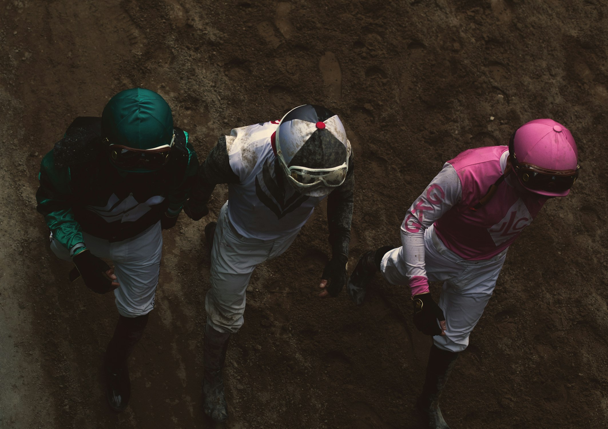 Eye-level, high-angle shot of three jockeys in racing gear, clad in vibrant pink, green, and white outfits, on a dirt track.