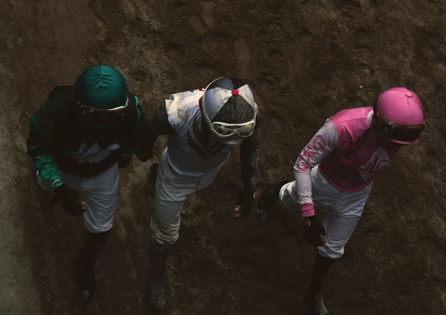 Eye-level, high-angle shot of three jockeys in racing gear, clad in vibrant pink, green, and white outfits, on a dirt track. 
