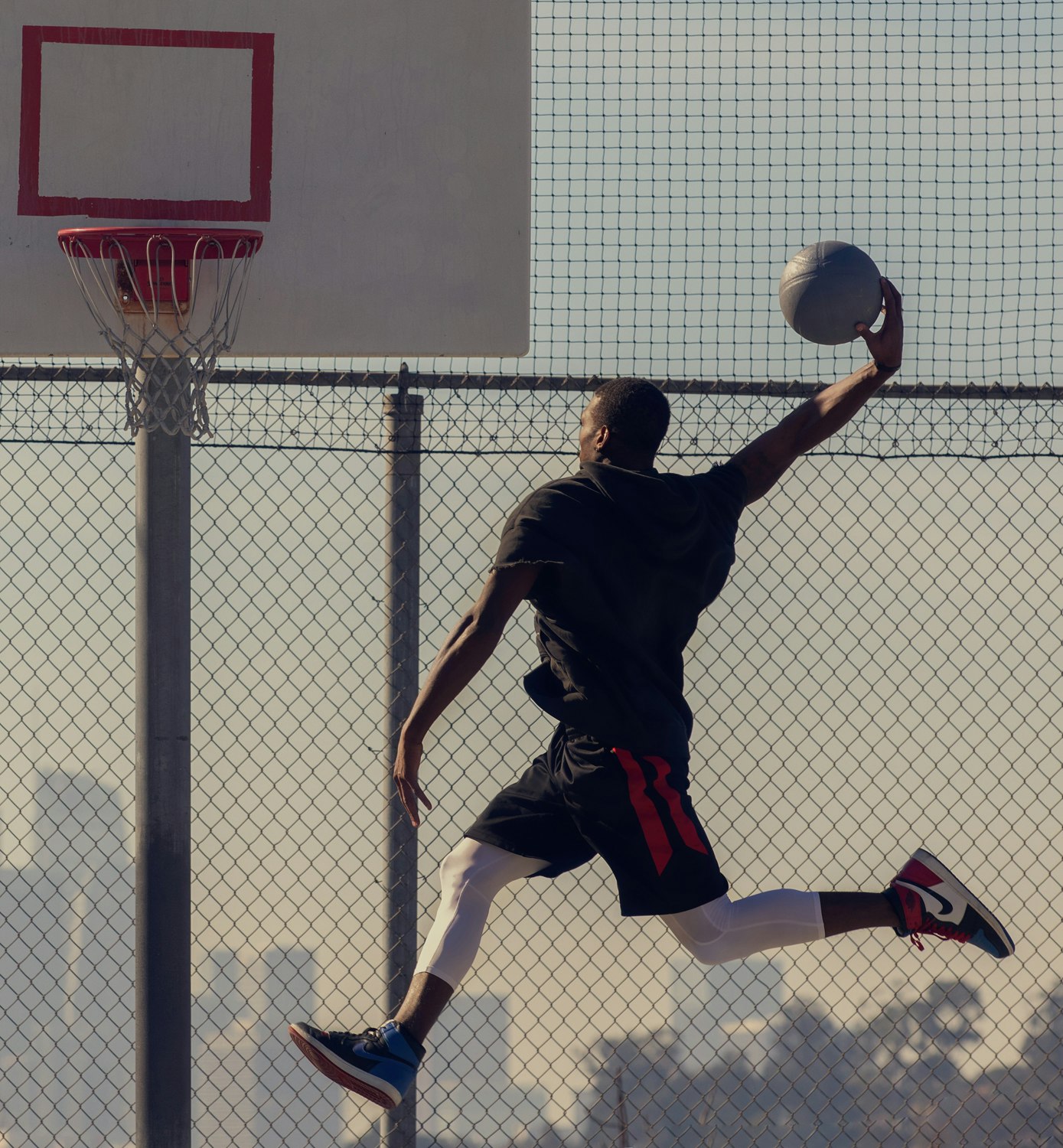 An African American man in athletic wear jumps to dunk a basketball on an outdoor court, captured in a dynamic, full shot against a blurred background.