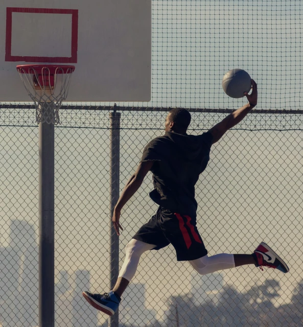An African American man in athletic wear jumps to dunk a basketball on an outdoor court, captured in a dynamic, full shot against a blurred background.