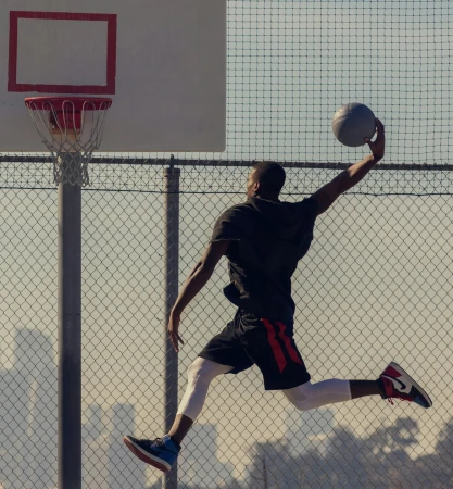 An African American man in athletic wear jumps to dunk a basketball on an outdoor court, captured in a dynamic, full shot against a blurred background.