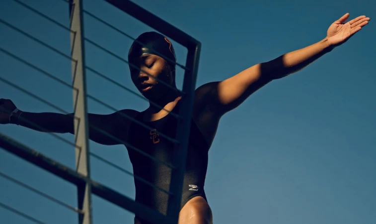 A young Black woman in a black swimsuit and swim cap stands in an athletic pose with arms outstretched against a bright blue sky and silver railing.