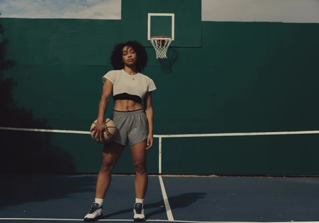 Athletic Black woman stands confidently on a blue basketball court holding a ball, with a green wall and hoop in the background.