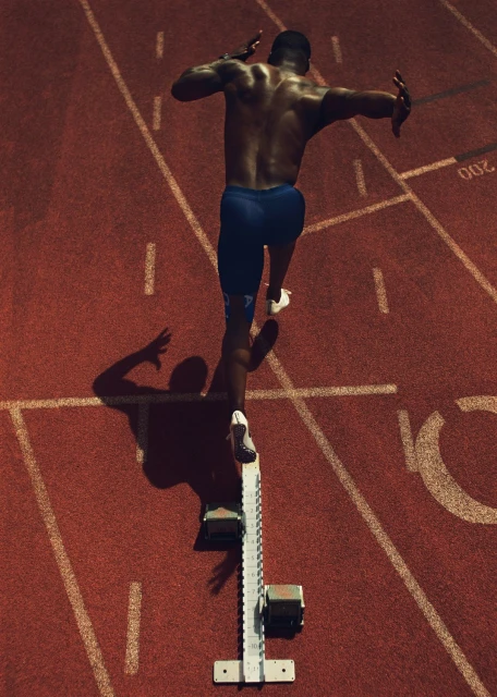 Overhead shot of a muscular adult male sprinter at the starting block on a red running track in blue shorts and white running shoes.