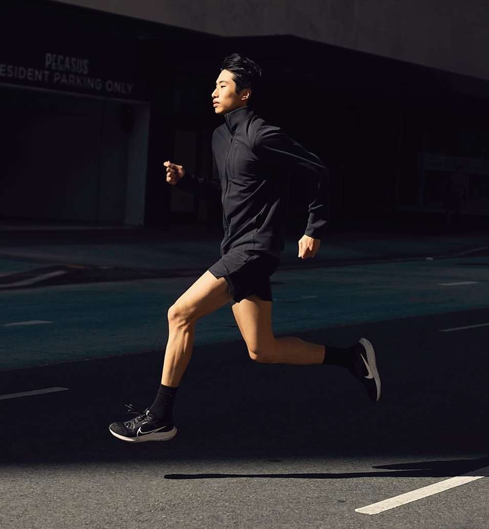 Asian man in dark athletic wear running outdoors on asphalt, bathed in light. Building and sign visible in the dark background.