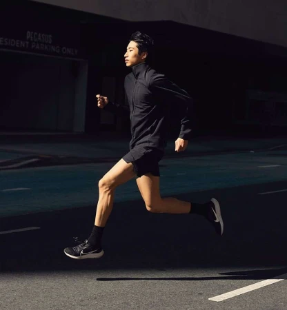 Asian man in dark athletic wear running outdoors on asphalt, bathed in light. Building and sign visible in the dark background.