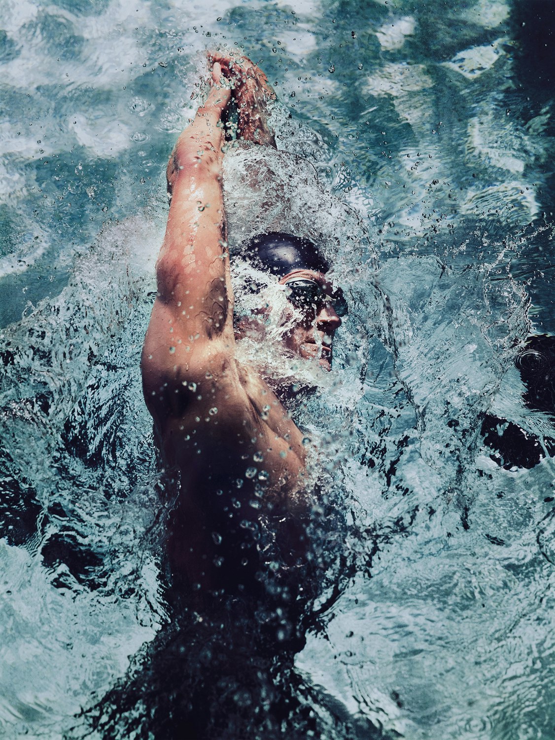 Underwater shot of an adult male swimmer in a black cap, with a competitive physique, swimming in a pool creating splashing water.