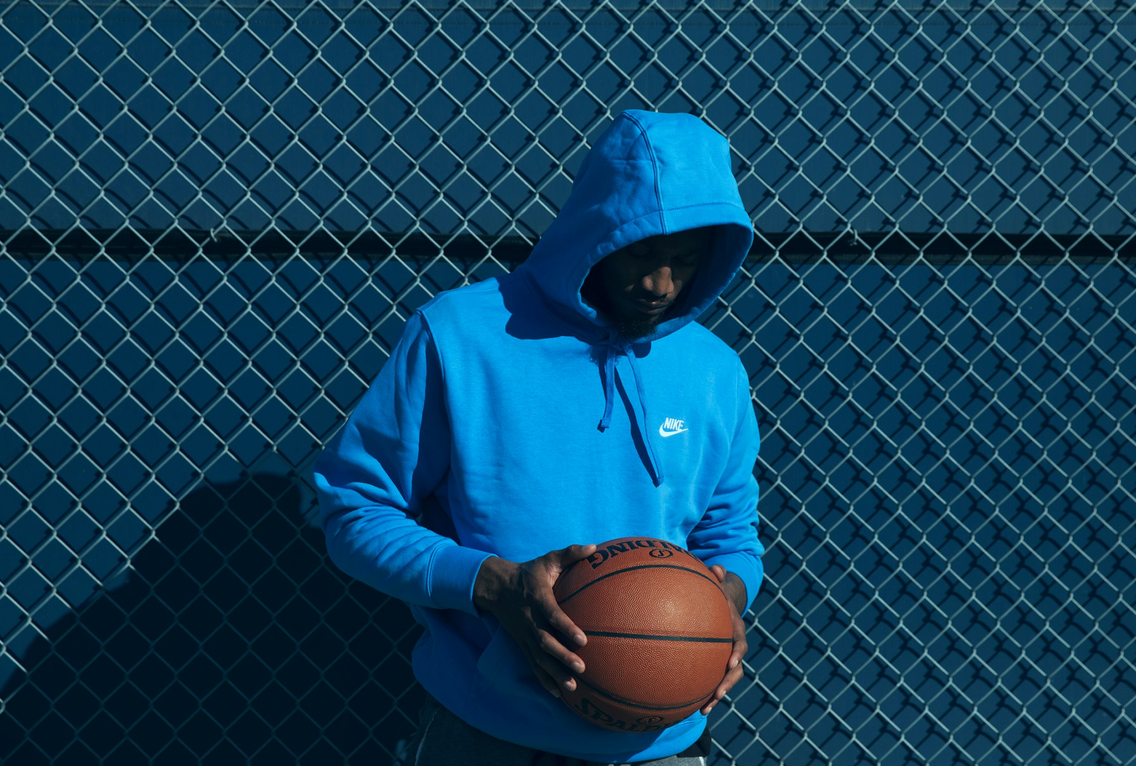 Man in blue hoodie holds a basketball in front of a blue chain-link fence, creating a sporty and casual aesthetic.