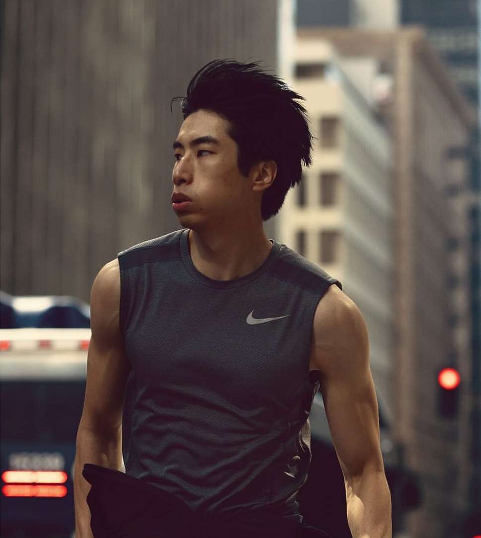 Adult male in a sleeveless athletic shirt poses outdoors in a city street with skyscrapers, looking to the side with a serious expression.