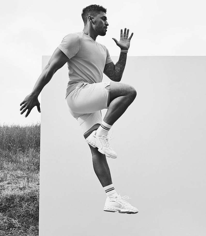 Athletic adult male in shorts and t-shirt leaps forward against a split backdrop of white and an outdoor field.