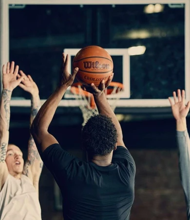 A man in a black shirt holds a basketball with other players and a basketball hoop in the background inside of an indoor court.