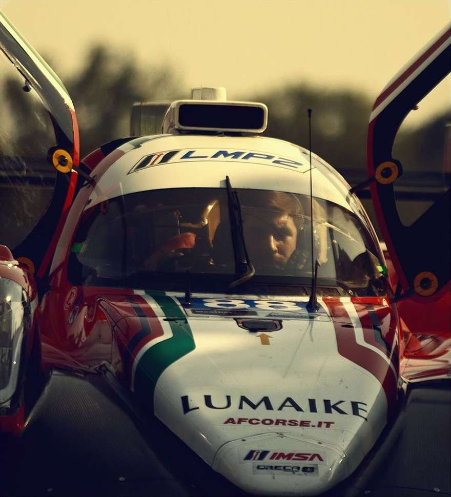 Close-up of a red, white, and green LMP2 race car with an adult in the driver's seat. The car has opened doors. The vehicle has branding.