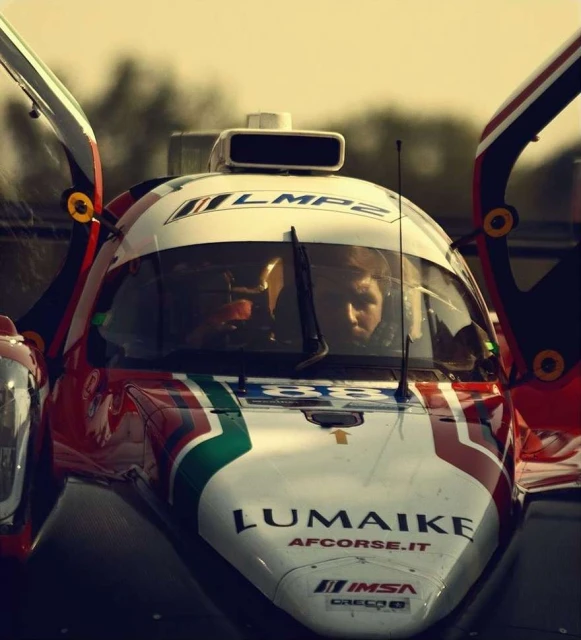 Close-up of a red, white, and green LMP2 race car with an adult in the driver's seat. The car has opened doors. The vehicle has branding.