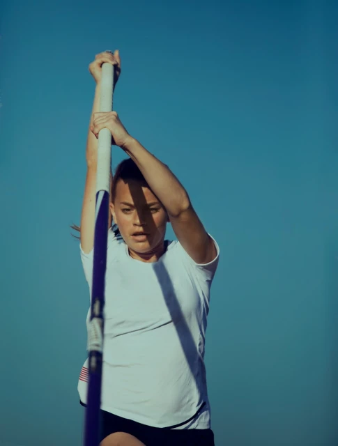Female pole vaulter grips the pole, preparing for her jump, against a clear blue sky on a sunny day during a sporting event.
