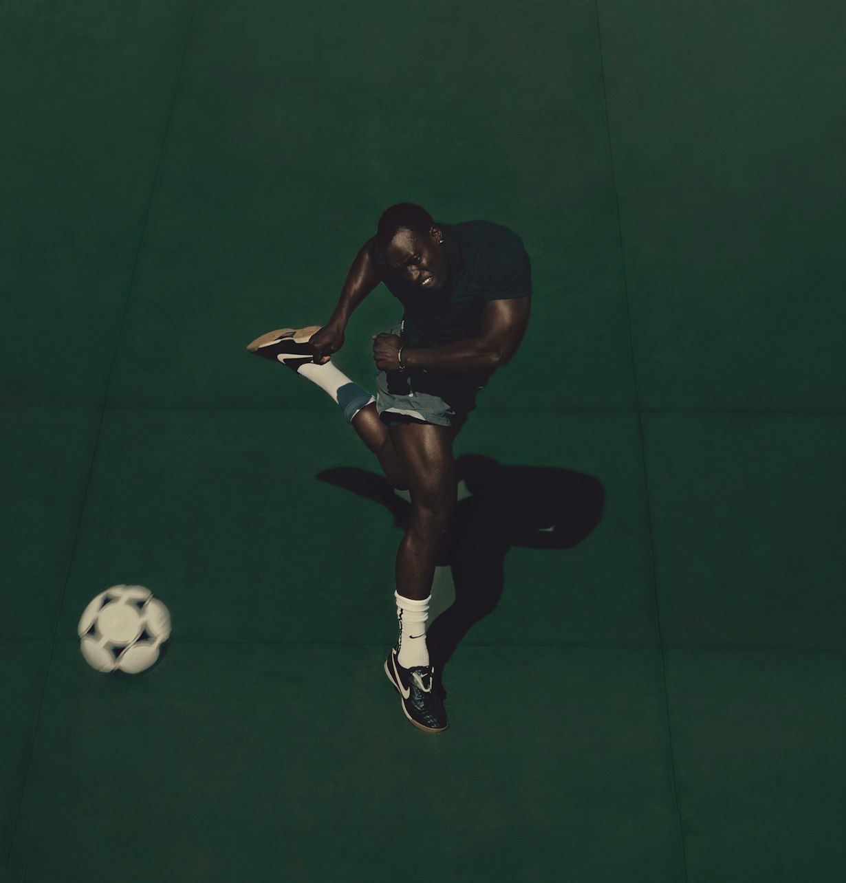 Overhead shot of an adult male kicking a soccer ball on a solid green background wearing a black shirt and grey shorts in a studio environment.