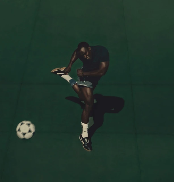 Overhead shot of an adult male kicking a soccer ball on a solid green background wearing a black shirt and grey shorts in a studio environment.