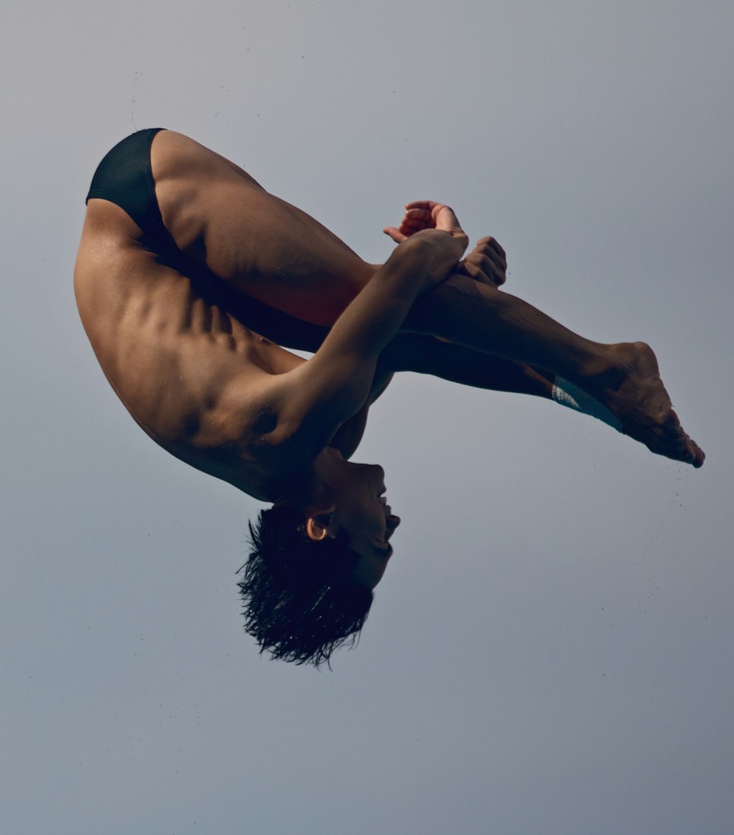 Male diver in mid-air performing a tuck, arms holding legs, set against a clear, light-blue sky. Athletic form and concentration.