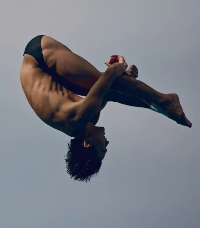 Male diver in mid-air performing a tuck, arms holding legs, set against a clear, light-blue sky. Athletic form and concentration.