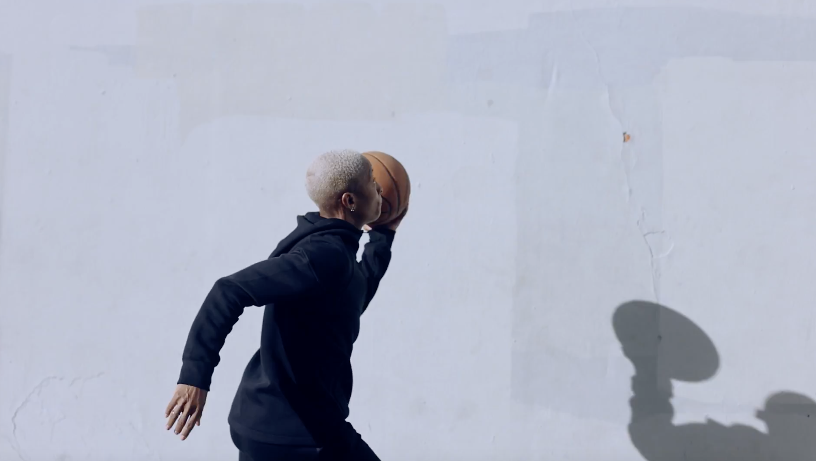 Short-haired person wearing a black hoodie running and holding a basketball against a plain white wall by Brian Lowe.