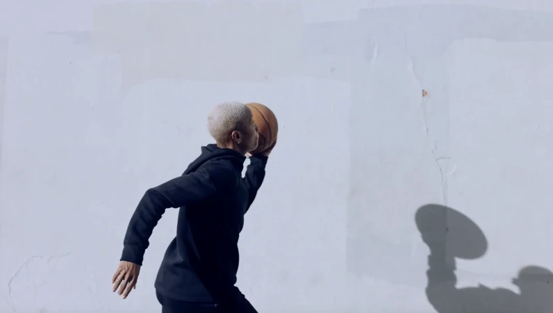 Short-haired person wearing a black hoodie running and holding a basketball against a plain white wall by Brian Lowe.