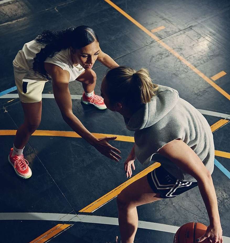 Two women play basketball indoors on a marked court, focusing on ball handling and teamwork, featuring athletic apparel and dynamic movement.
