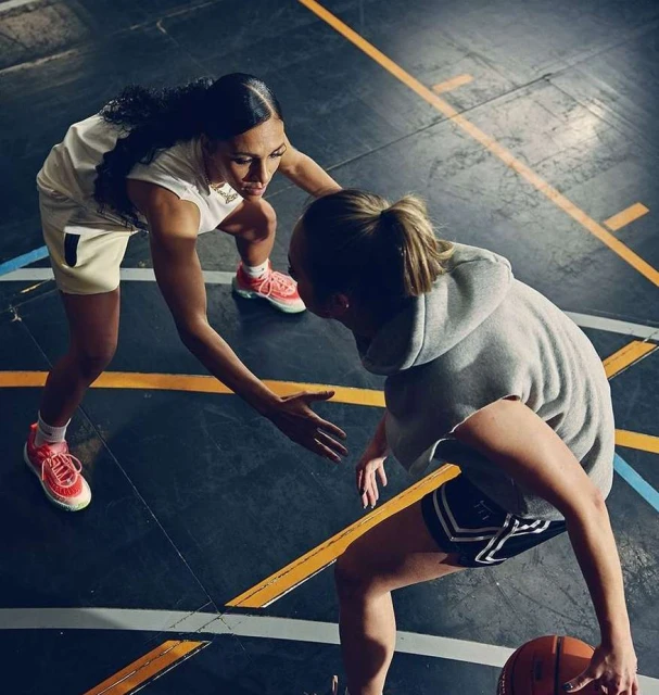 Two women play basketball indoors on a marked court, focusing on ball handling and teamwork, featuring athletic apparel and dynamic movement.