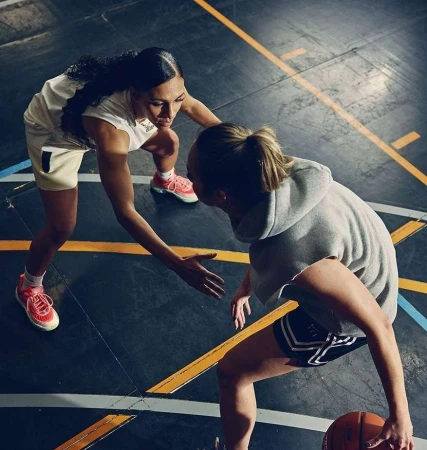 Two women play basketball indoors on a marked court, focusing on ball handling and teamwork, featuring athletic apparel and dynamic movement.