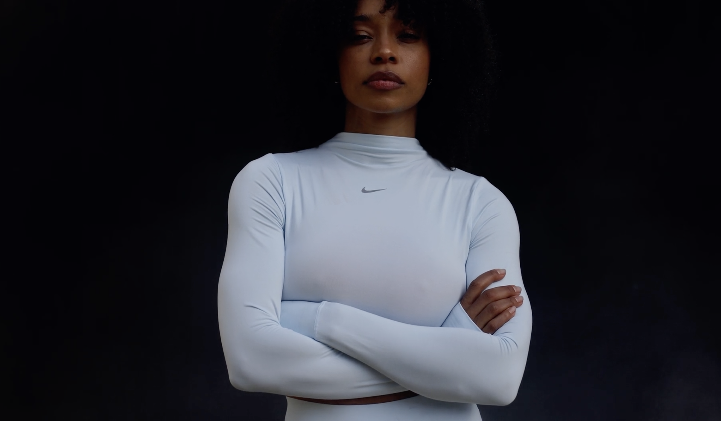 Athletic woman with arms crossed in light blue long-sleeve activewear top in a studio, showcasing sportswear fashion against dark backdrop.