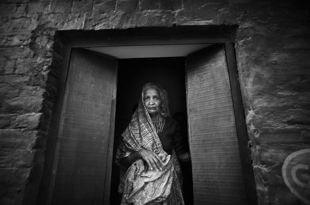 Monochrome portrait of an elderly woman in a patterned shawl standing in a doorway with aged brick and wooden doorframe.