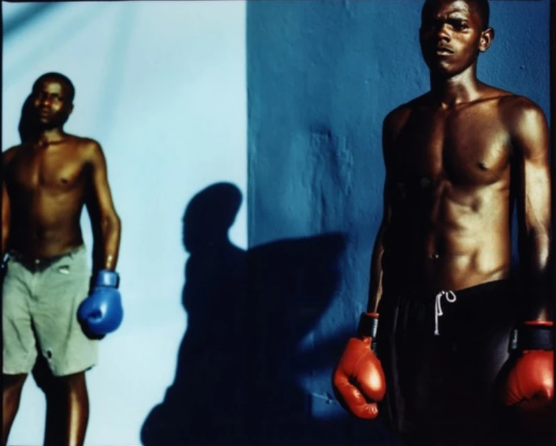 Two muscular shirtless Black male boxers with boxing gloves pose against a blue and white wall in a studio setting with dramatic lighting.