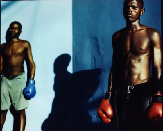 Two muscular shirtless Black male boxers with boxing gloves pose against a blue and white wall in a studio setting with dramatic lighting.