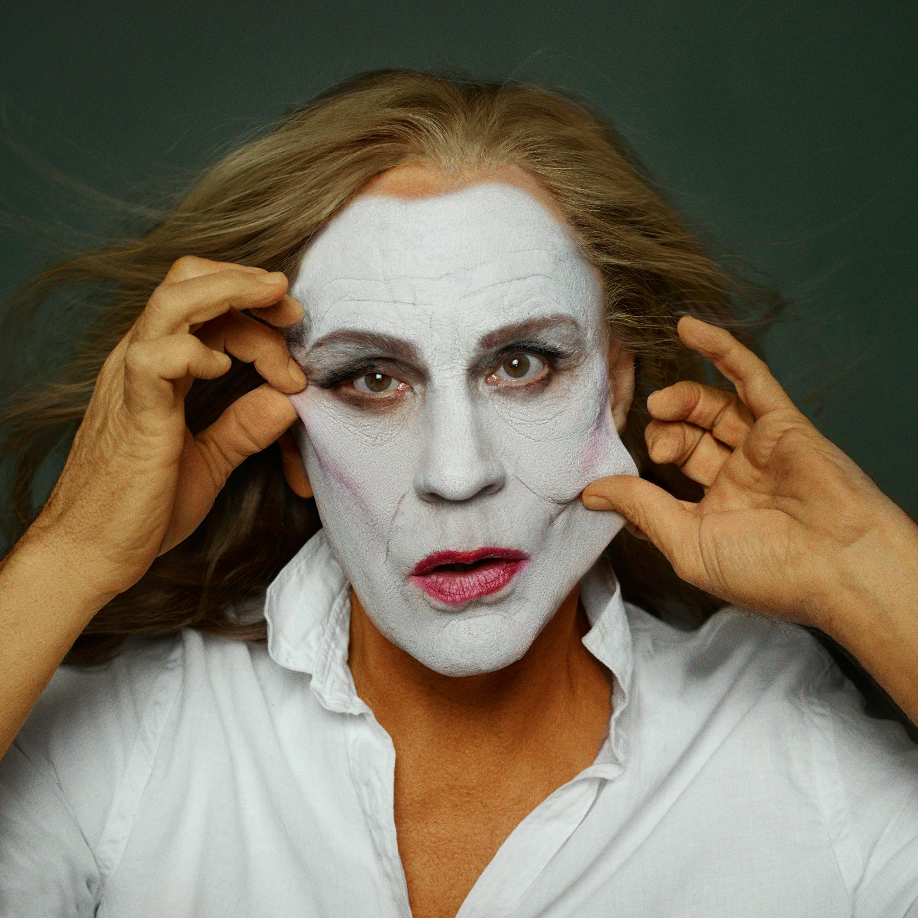 Portrait of a person with white makeup holding up their face, wearing a white shirt against a dark green background in a studio.