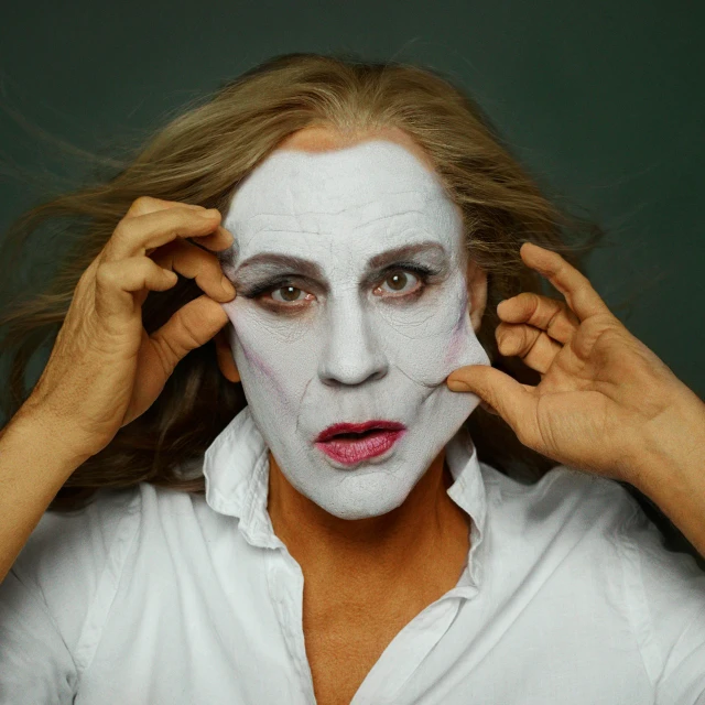 Portrait of a person with white makeup holding up their face, wearing a white shirt against a dark green background in a studio.